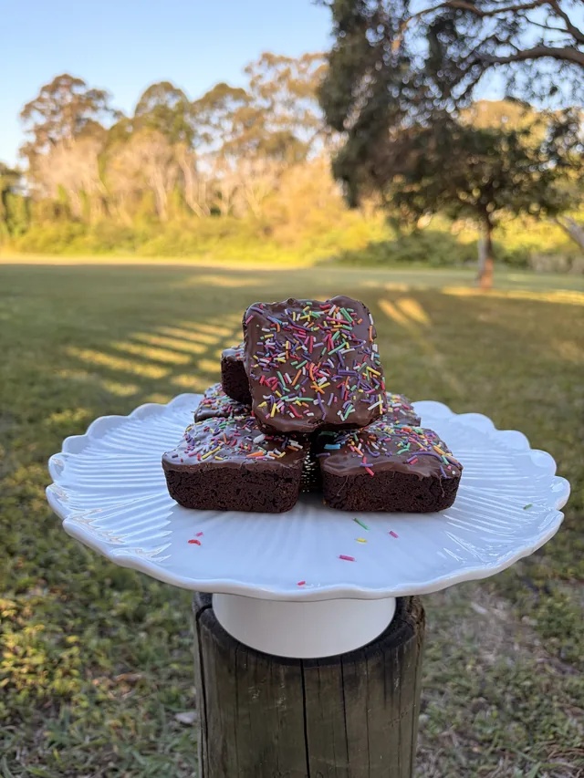 A stack of glazed Fudgy Brownies with colourful sprinkles on a cake stand outdoors.
