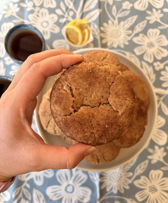 A close-up Cinnamon Hug cookie held above a plate of cookies and tea.