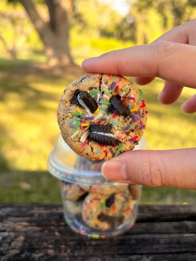 A close-up Happy Cookie Yummy Bite with Oreo pieces and rainbow sprinkles held outdoors.